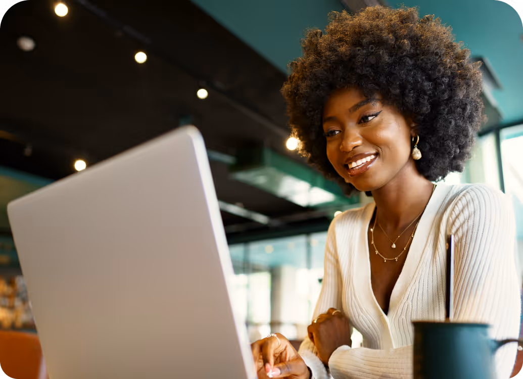 Smiling woman with curly hair using laptop in modern indoor space with a mug nearby.