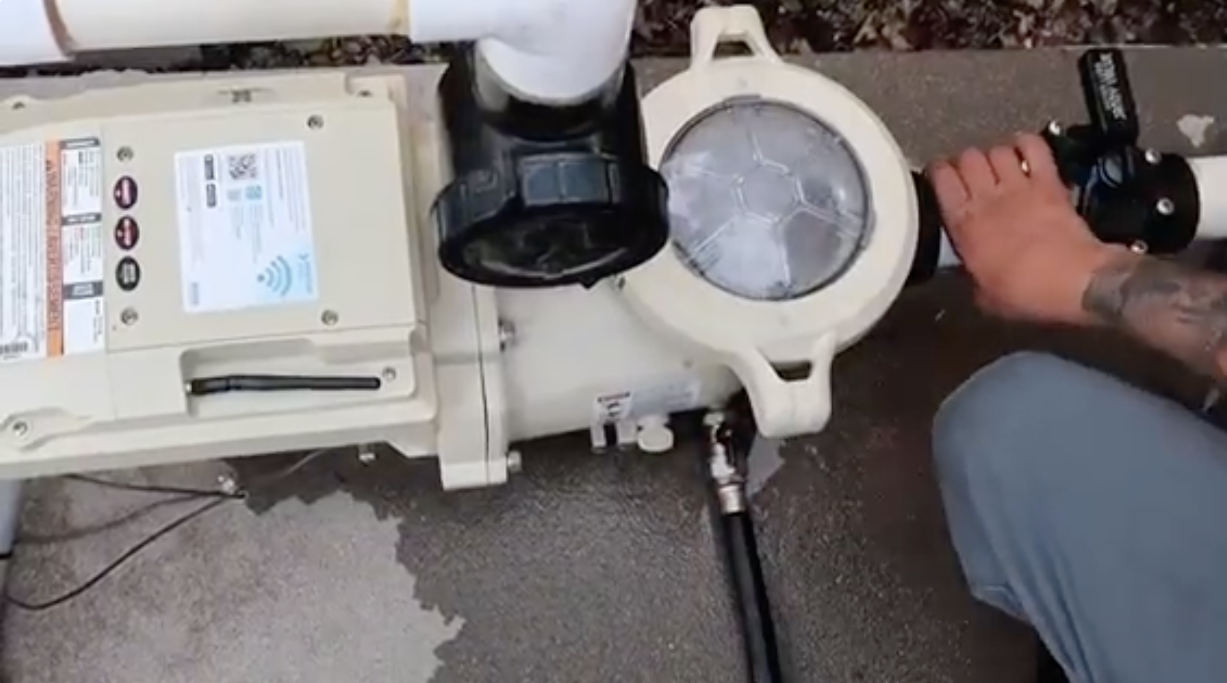 Hand with tattoo adjusting a valve on a beige pool pump with connected white pipes.