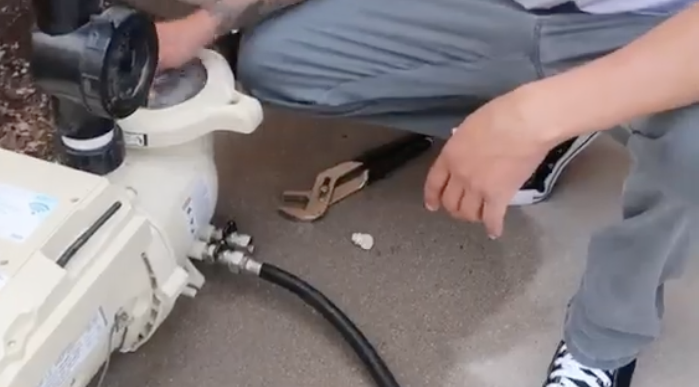 Person adjusting a pool pump with tools while kneeling on the ground near plumbing pipes.