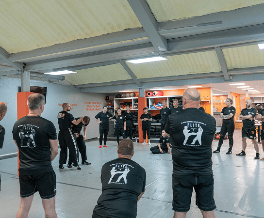Group of people in black martial arts uniforms standing on mats in a gym during a training session.