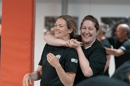 Two women smiling while practicing self-defense or martial arts in a gym setting.