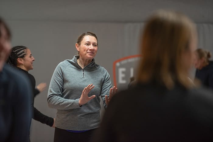 Woman in gray pullover speaking and gesturing with hands among a small group in an indoor setting.
