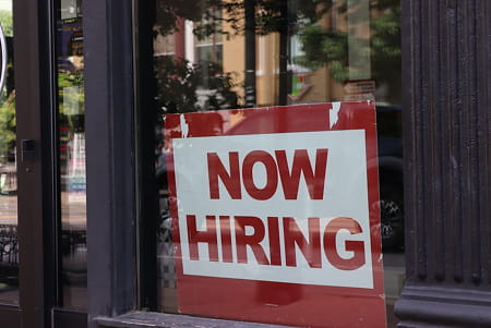 Red and white sign on a window displaying the text NOW HIRING.