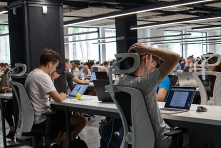 People working on laptops at desks in a modern open office space with large windows.