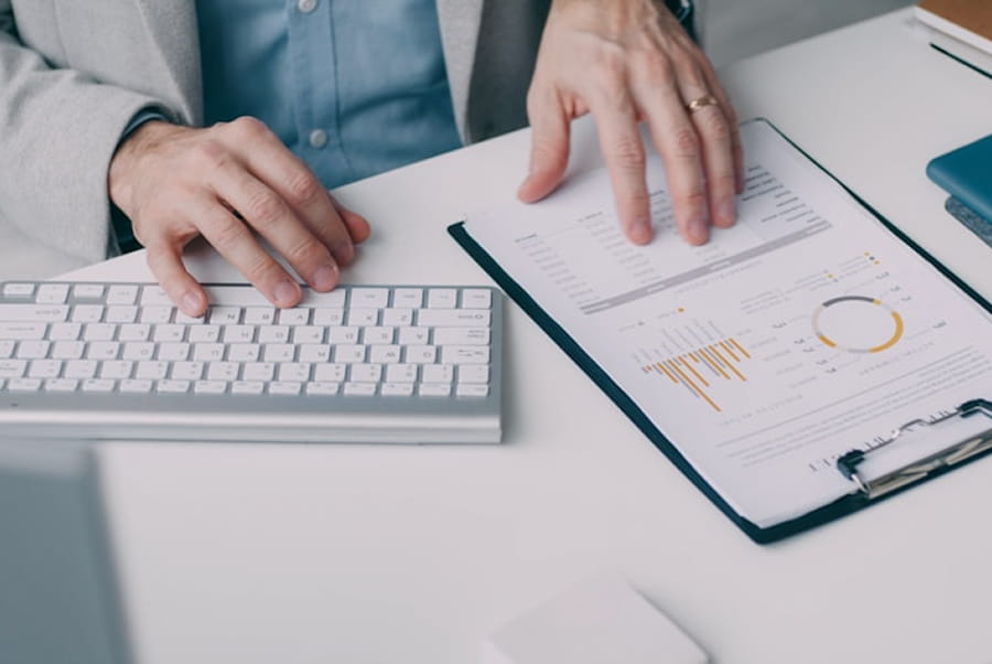 Hands working with a keyboard and financial charts on a clipboard at a white desk.