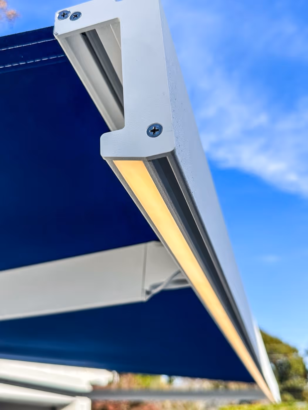 Close-up of white metal frame with warm LED strip light under a blue outdoor awning against a blue sky.