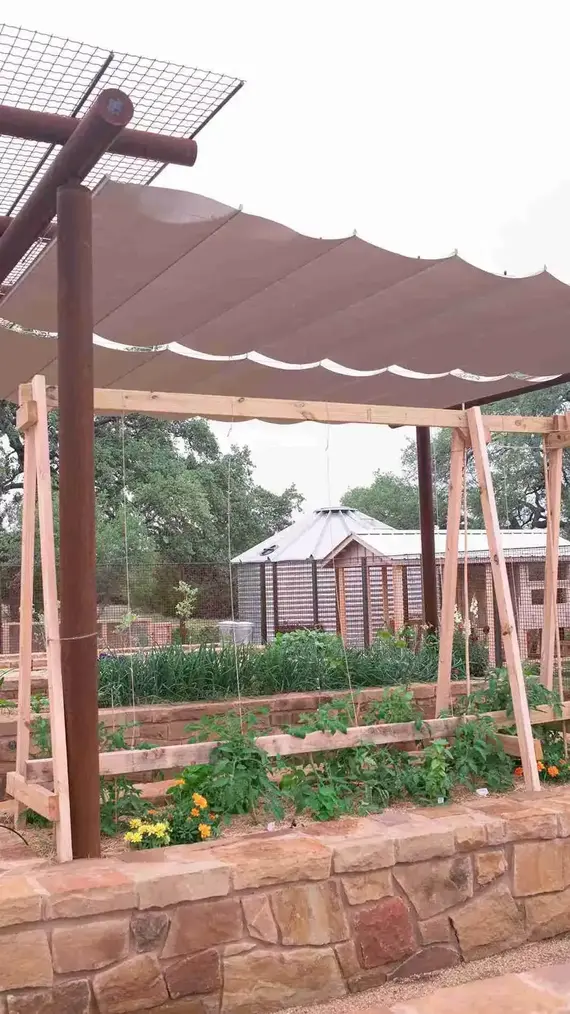 Raised garden beds with plants under a shade cloth supported by wooden frame and stone wall.