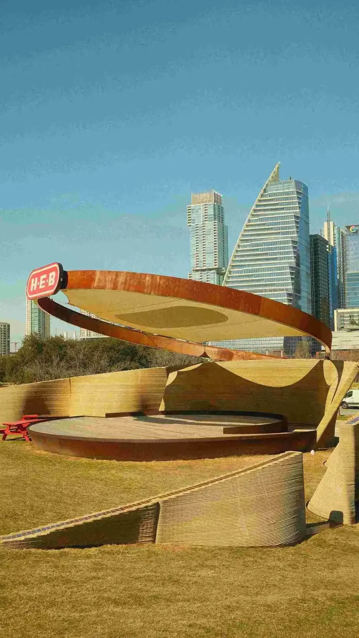 Large outdoor stage with curved roof labeled H-E-B in a city park with skyscrapers in background.