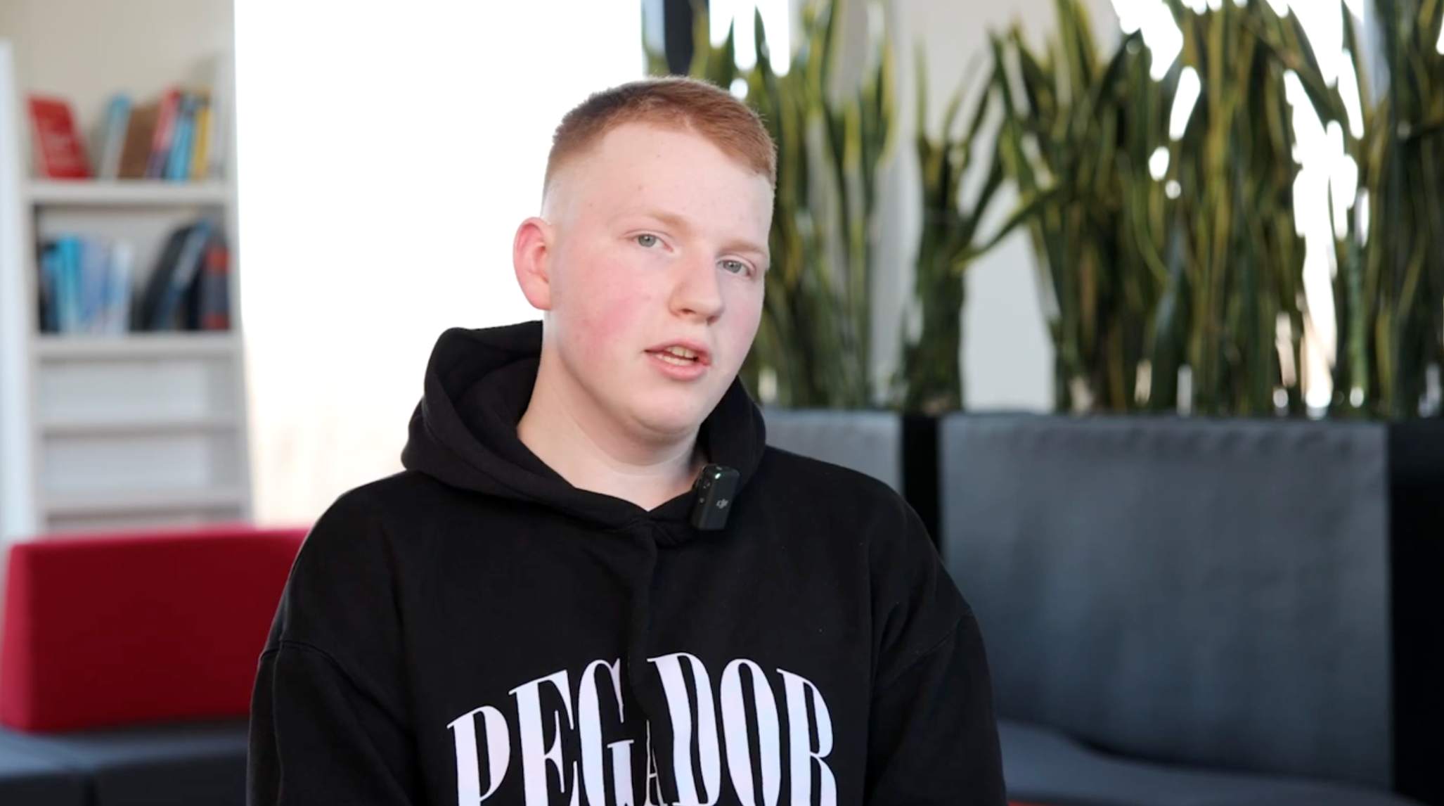 Junger Mann mit kurzem roten Haar trägt schwarzen Hoodie mit der Aufschrift PEGADOR und sitzt in einem modernen Raum mit Pflanzen und Bücherregal.