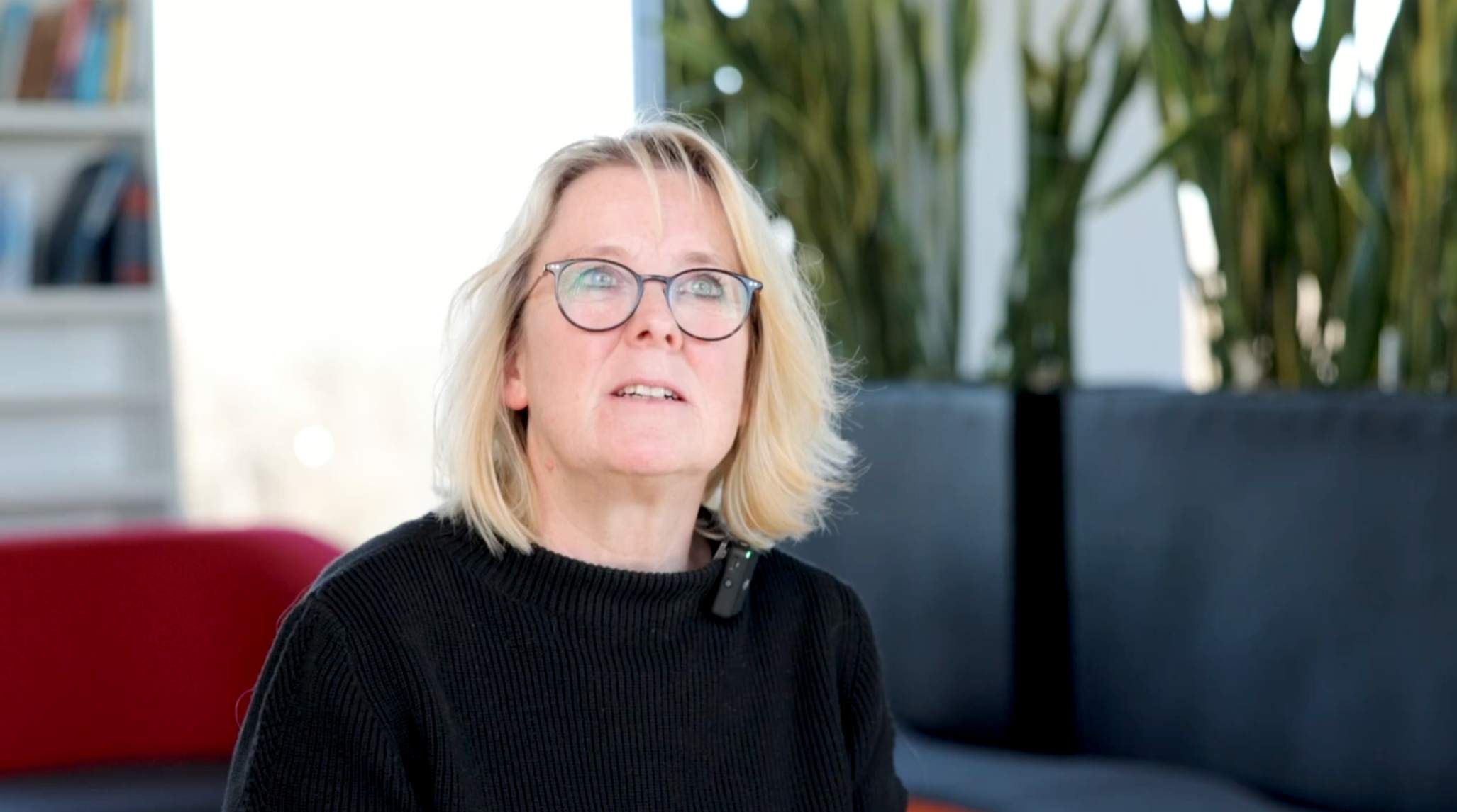 Blonde Frau mit Brille und schwarzem Pullover sitzt in einem modernen Raum mit Pflanzen und einem roten Sofa im Hintergrund.
