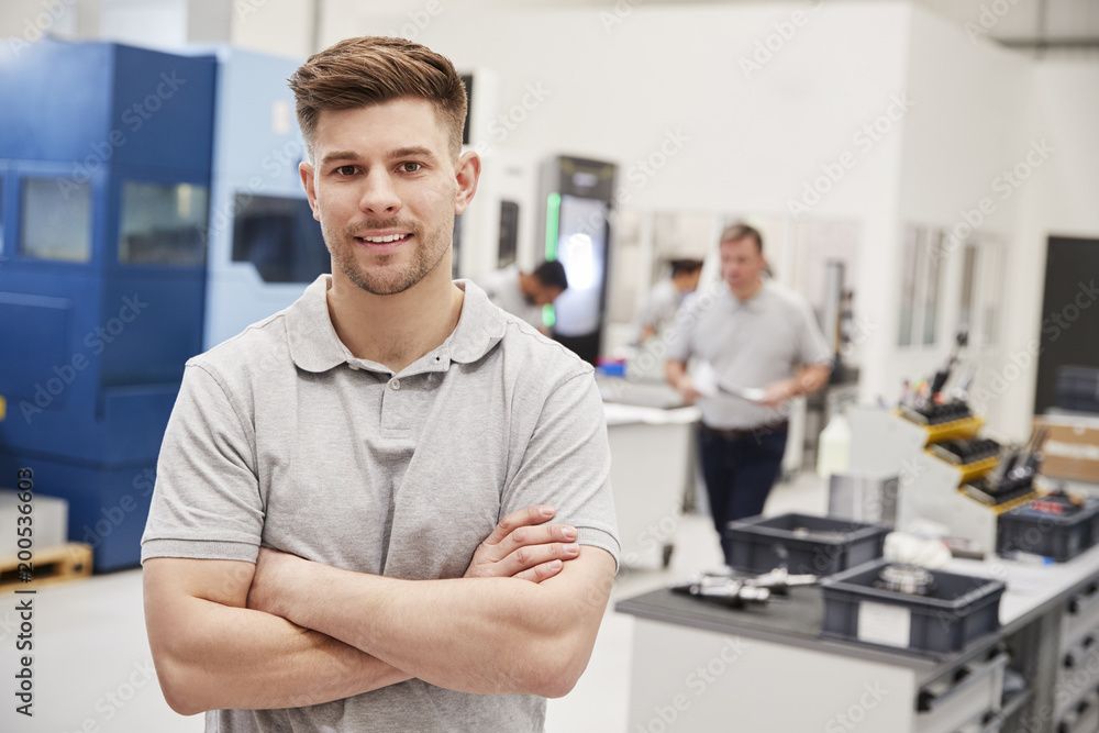 Junger Mann in einem grauen Poloshirt steht mit verschränkten Armen in einer Fabrik oder Werkstatt.