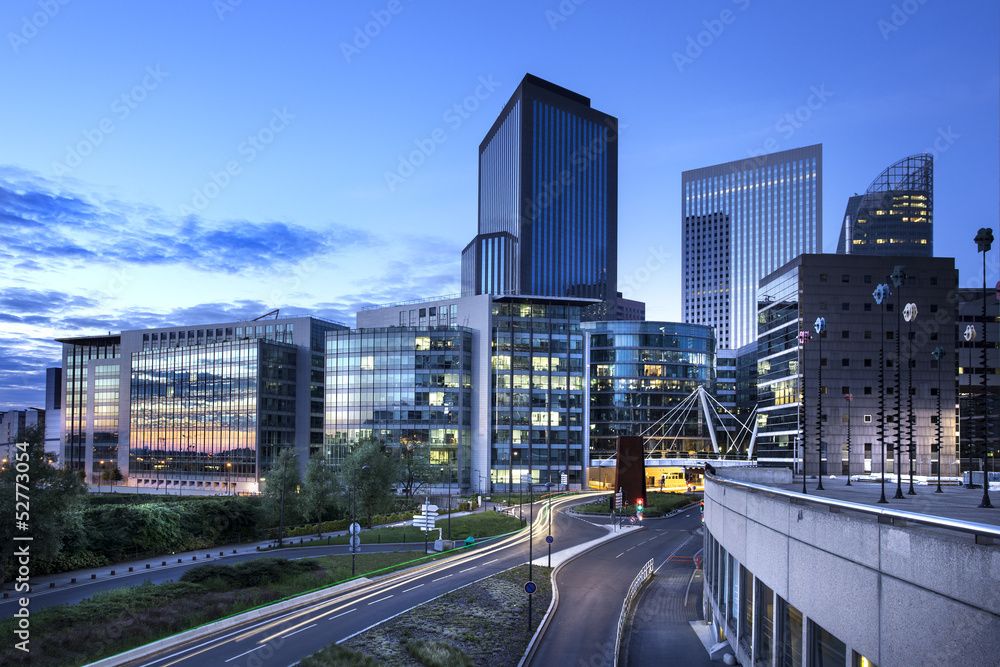 Moderne Bürogebäude mit Glasfassaden und beleuchteten Fenstern bei Dämmerung in einer Stadtlandschaft.