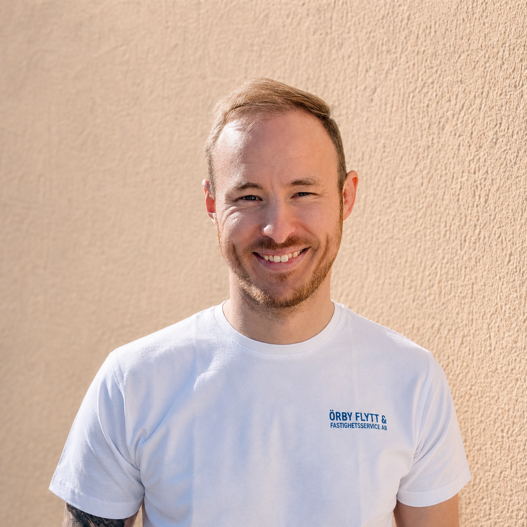 Smiling man with short light brown hair wearing a white t-shirt with blue text standing against a beige textured wall.