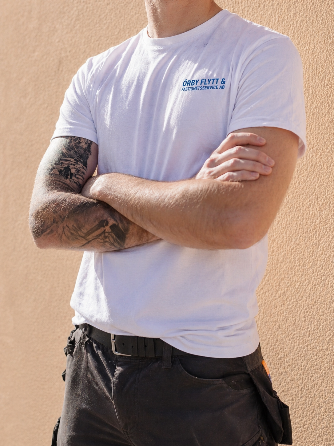 Person wearing a white t-shirt with blue text and black pants, arms crossed, showing a tattoo on the left forearm.