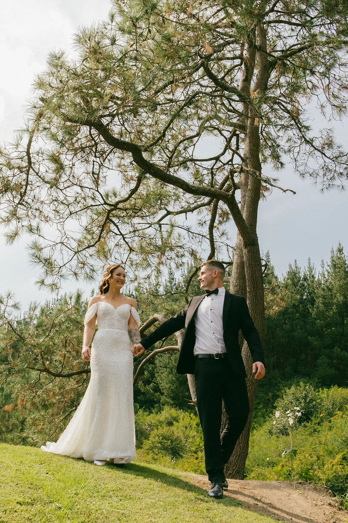 Bride in white wedding dress and groom in black suit holding hands outdoors near pine trees.