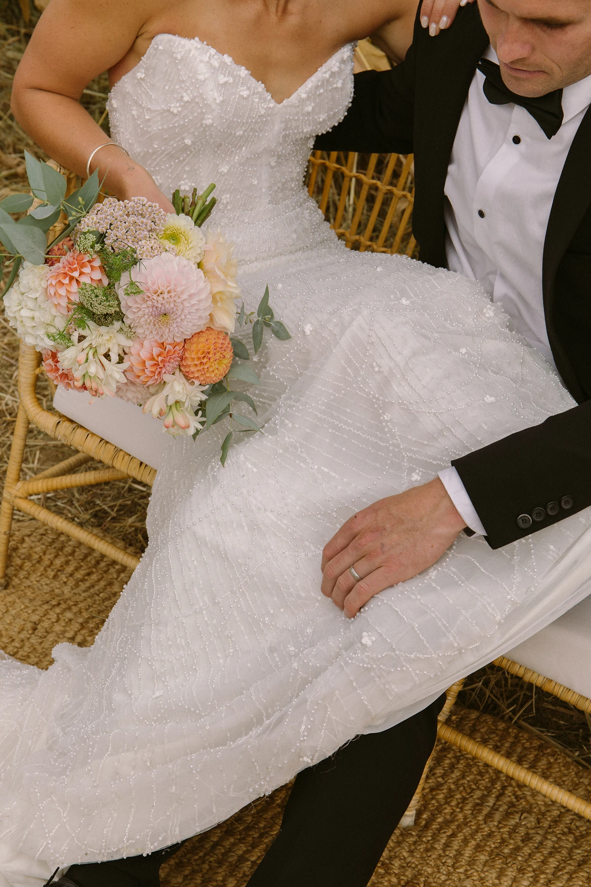 Bride in beaded white gown holding pastel flower bouquet sitting next to groom in black tuxedo.