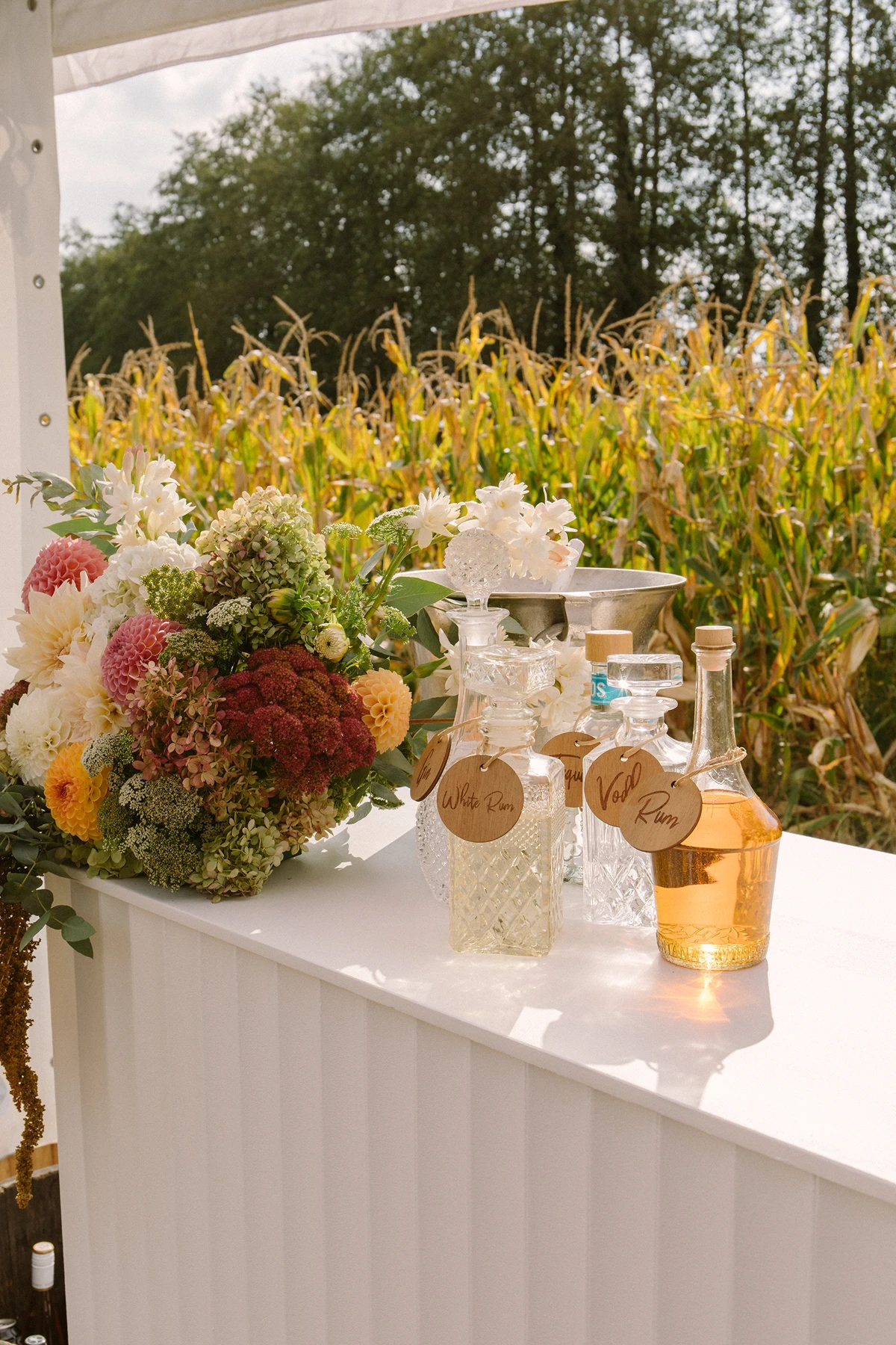 Flower bouquet and glass bottles labeled White Rum and Vodka on a white bar counter outside by cornfield.
