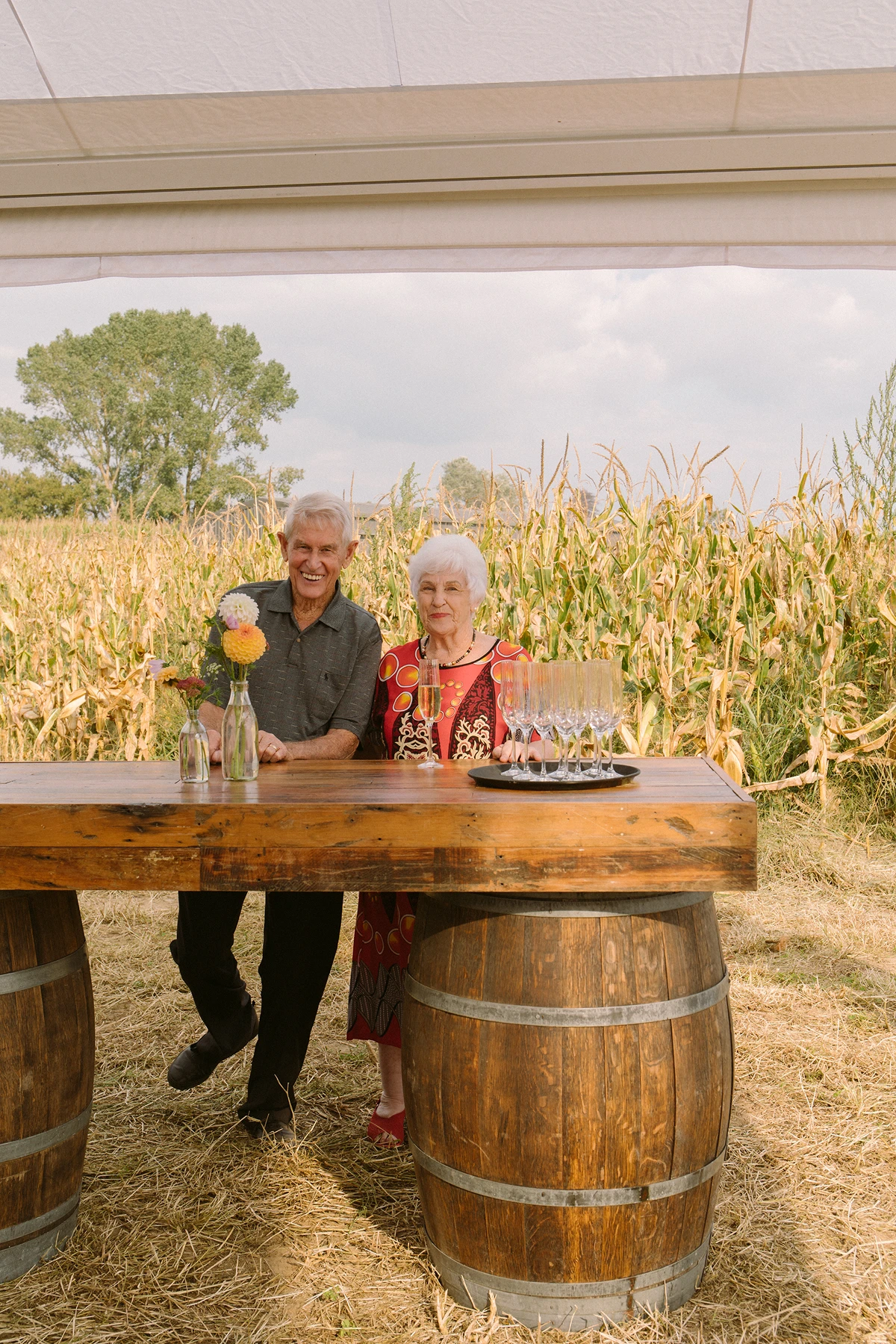 Elderly man and woman smiling behind wooden bar with flowers and champagne glasses outdoors.