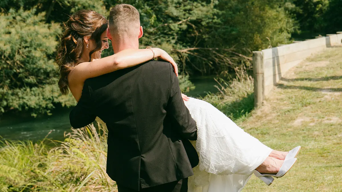 Groom in black suit carrying smiling bride in white dress outdoors near a river and grass.