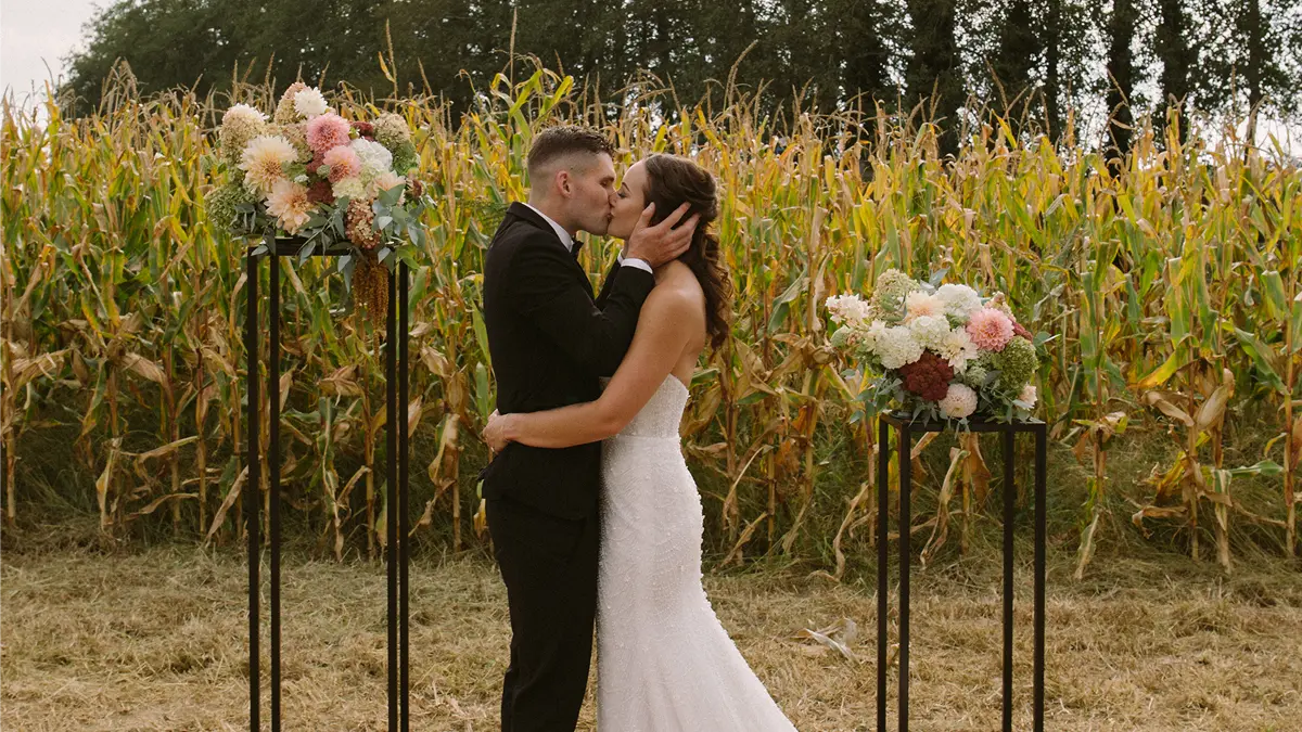 Bride and groom kissing in front of a cornfield with floral arrangements on black stands beside them.