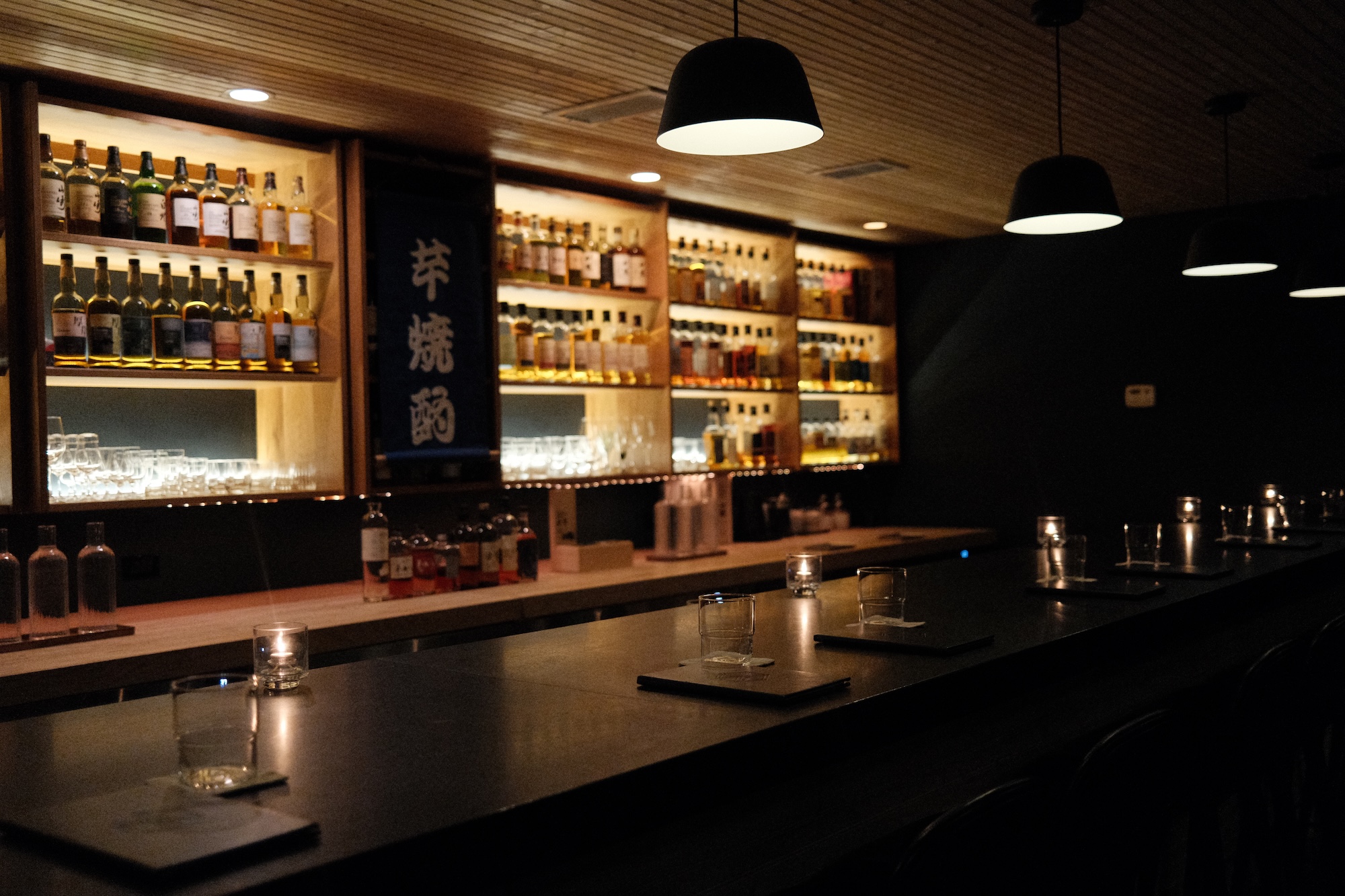 The Kumiko whisky bar counter, backlit shelves of Japanese whisky and a navy shōchū banner behind
