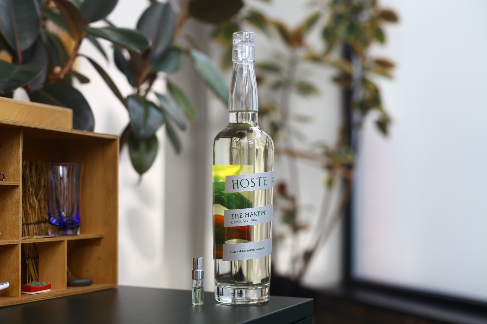 A clear 750ml Hoste bottle of The Martini with a yuzu atomizer beside it, on a dark counter