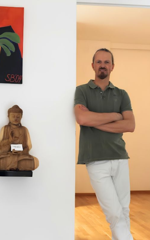 Man with a beard and mustache wearing a green shirt and white pants leaning against a wall inside a room.