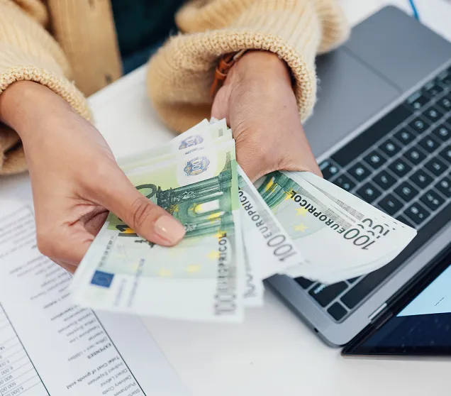 Person zählt mehrere 100-Euro-Scheine über einem Laptop auf einem weißen Tisch.