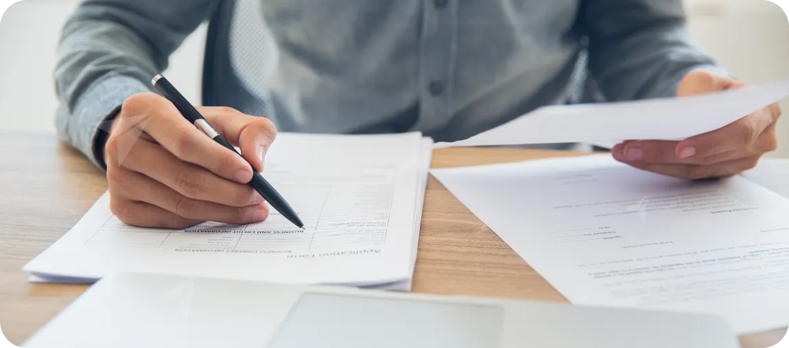 Person, die ein Formular mit einem Stift auf einem Holztisch ausfüllt und dabei ein Blatt Papier hält.