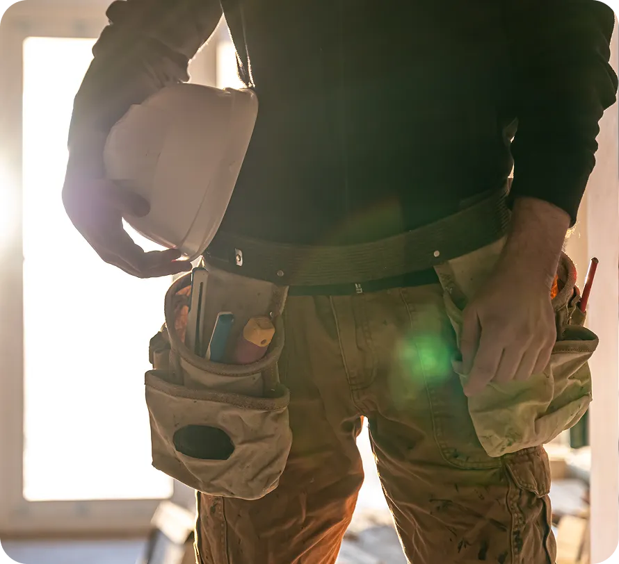 Handwerker hält einen weißen Schutzhelm und trägt einen Werkzeuggürtel mit mehreren Werkzeugen.