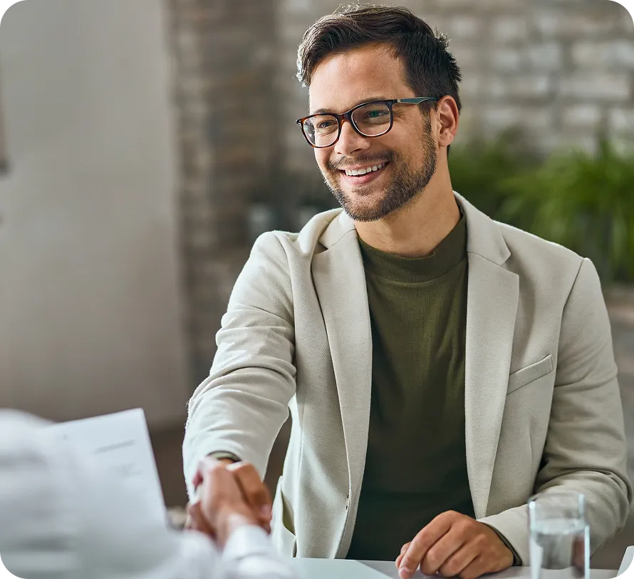 Lächelnder Mann mit Brille und beige Blazer reicht die Hand zum Handschlag über einen Tisch.