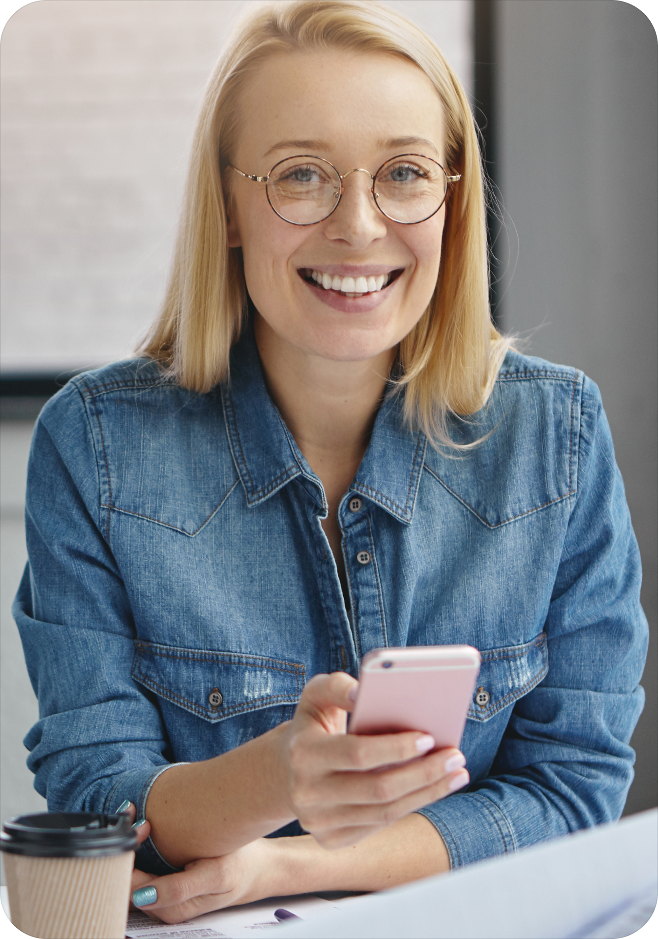 Lächelnde Frau mit blonden Haaren und runder Brille hält ein rosa Smartphone und trägt eine Jeansjacke.