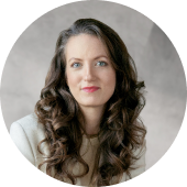 Portrait of a woman with long, curly brown hair wearing a light-colored jacket and a neutral top, against a softly blurred gray background.