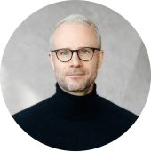 Portrait of a middle-aged man with short gray hair and beard, wearing black round glasses and a black turtleneck sweater, against a gray textured background.
