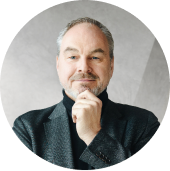 Middle-aged man with gray hair and beard wearing a dark jacket, resting his chin on his hand and looking at the camera, against a neutral light gray background.