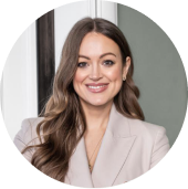 Portrait of a smiling woman with long brown hair wearing a light beige blazer and gold necklace, sitting indoors.