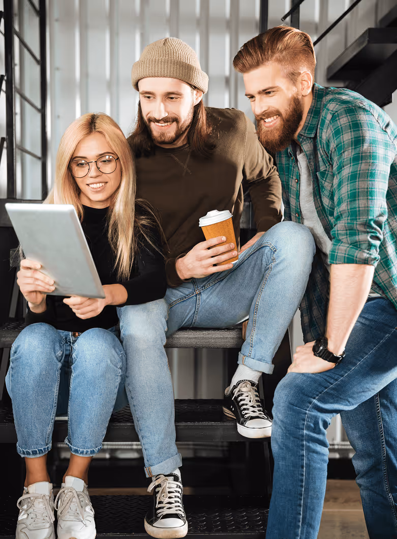 Three young colleagues sitting on stairs smiling and looking at a tablet, one holding a coffee cup.