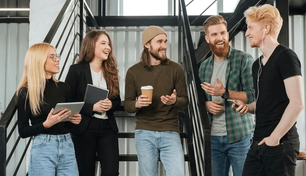 Young colleagues smiling on black stairs, one holding coffee, one with a tablet.