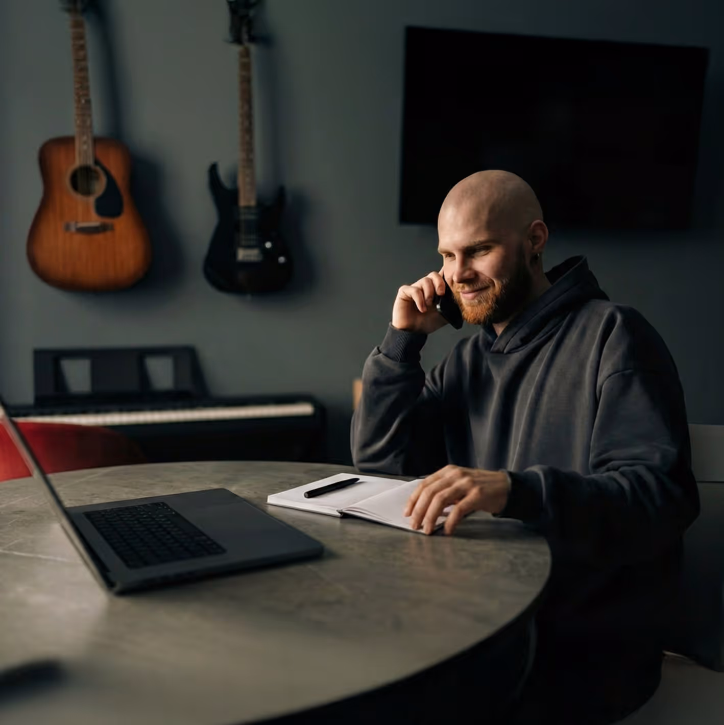 Smiling bald man sitting at a round table with a laptop and notebook, talking on a phone in a room with guitars and a keyboard.