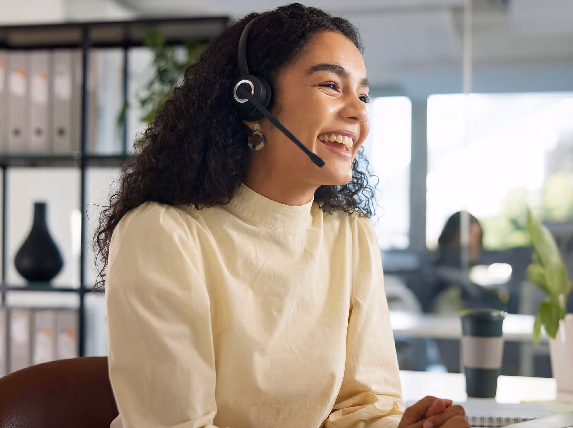 Smiling woman with curly hair wearing a headset, working in a bright office environment.