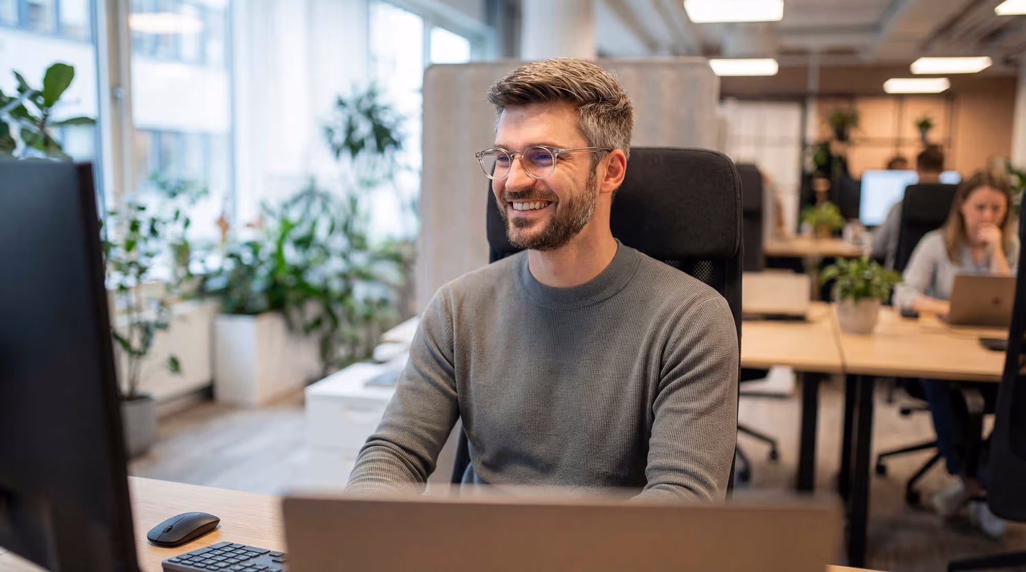 Smiling man with glasses sitting at a desk in a modern office with plants and colleagues in the background.