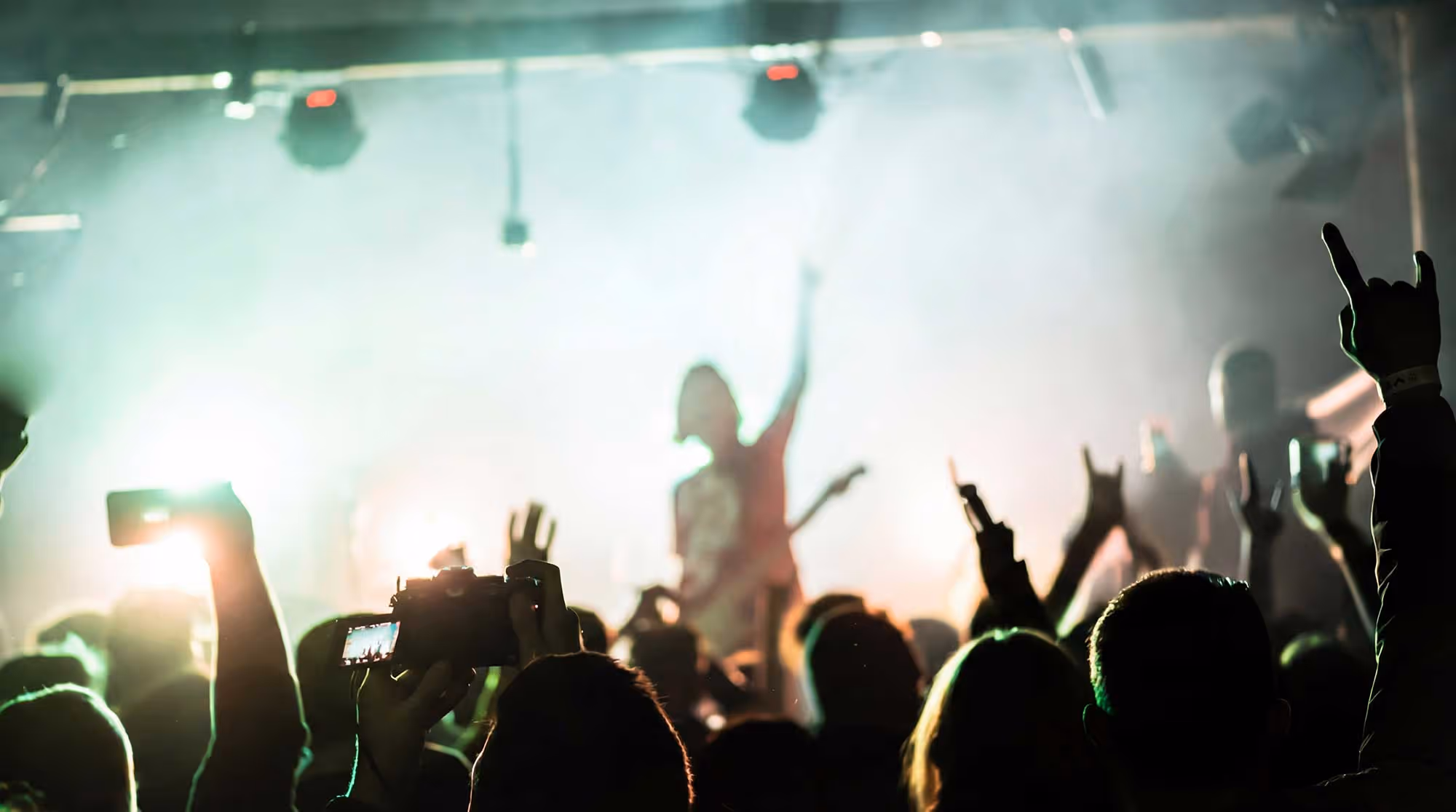 Crowd at a concert with raised hands and one person holding a camera, performer blurred in the bright stage lights.