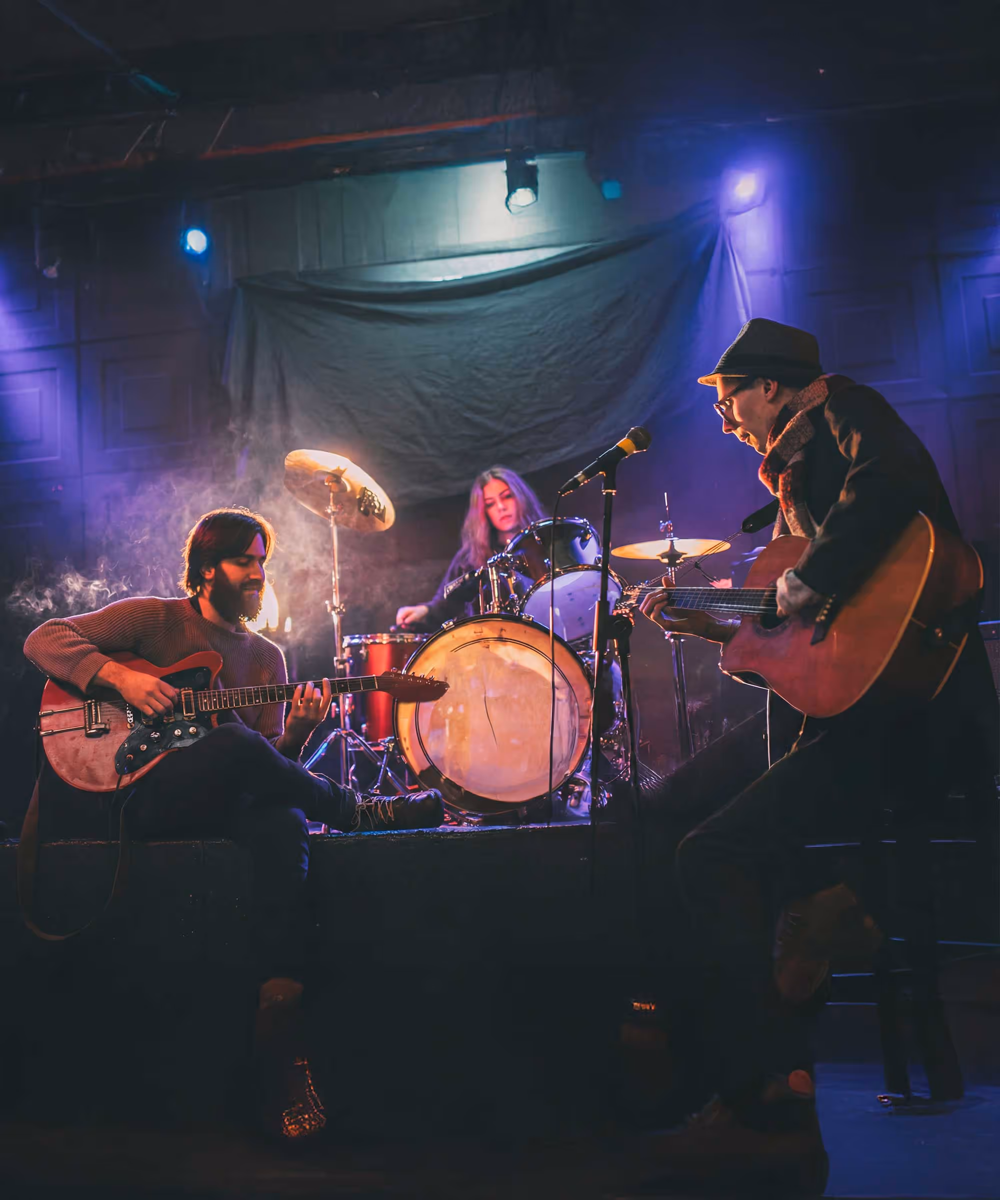 Three musicians performing on stage with electric guitar, acoustic guitar, and drum set under blue and purple lighting.