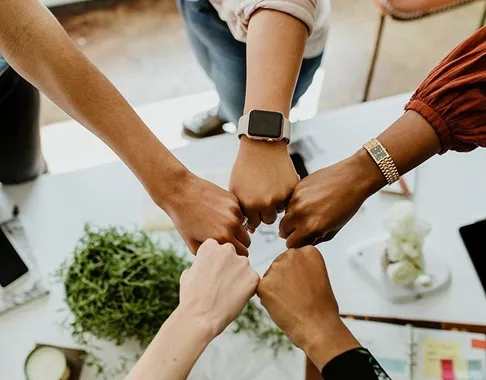 Five diverse hands joined together in a fist bump over a desk with office supplies and a small plant.
