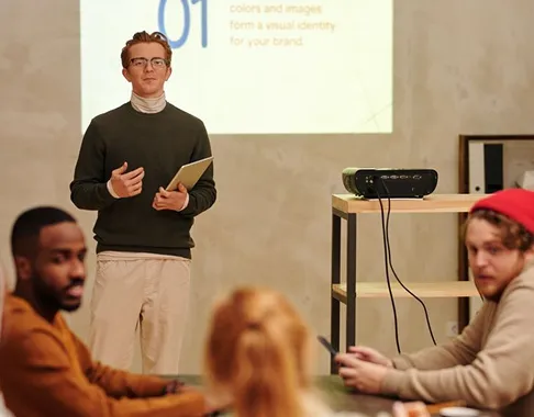 Man with glasses holding a tablet presenting to a group with a projector and screen in the background.