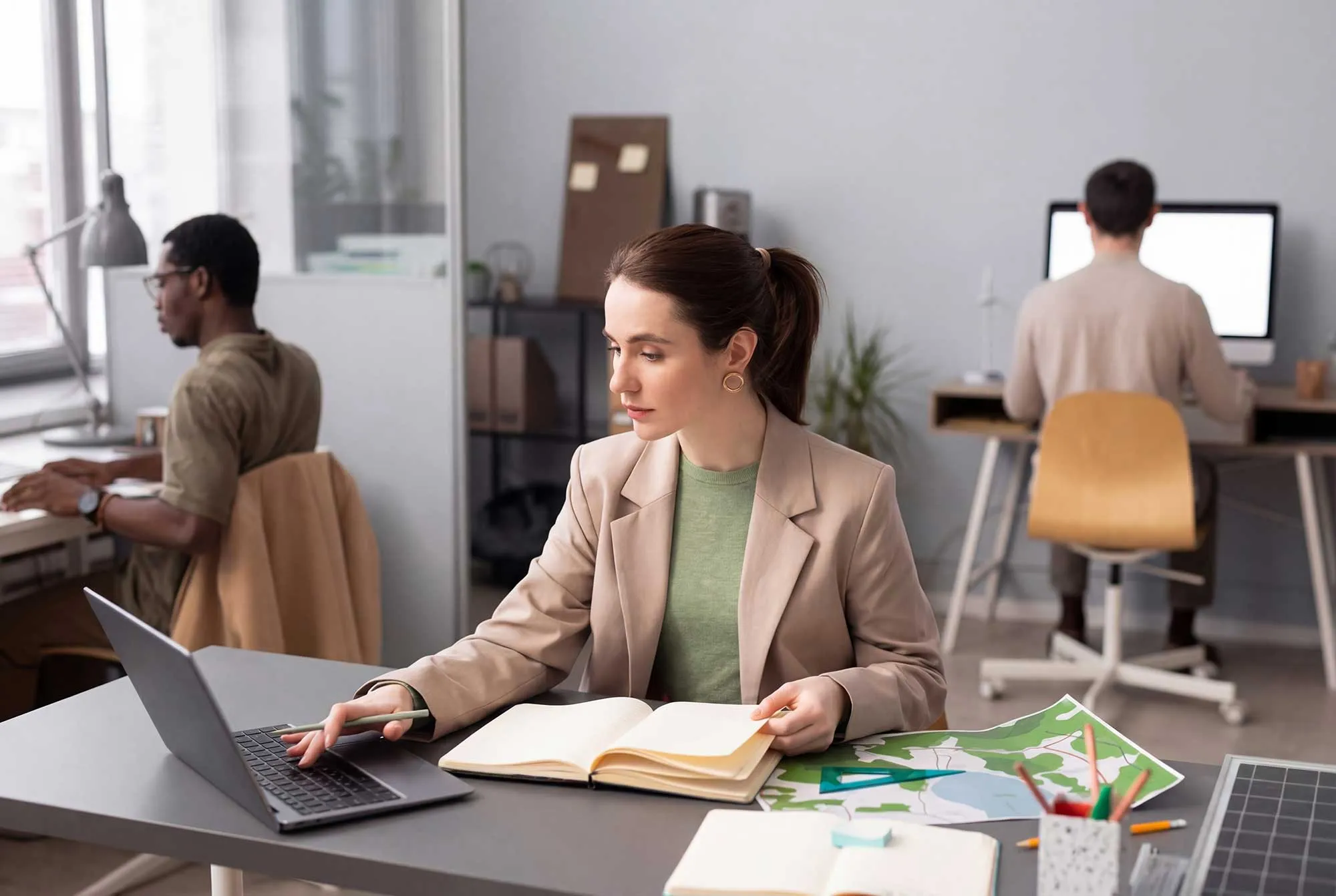 Woman in a beige blazer working at a desk with a laptop, open book, and maps in a shared office space.
