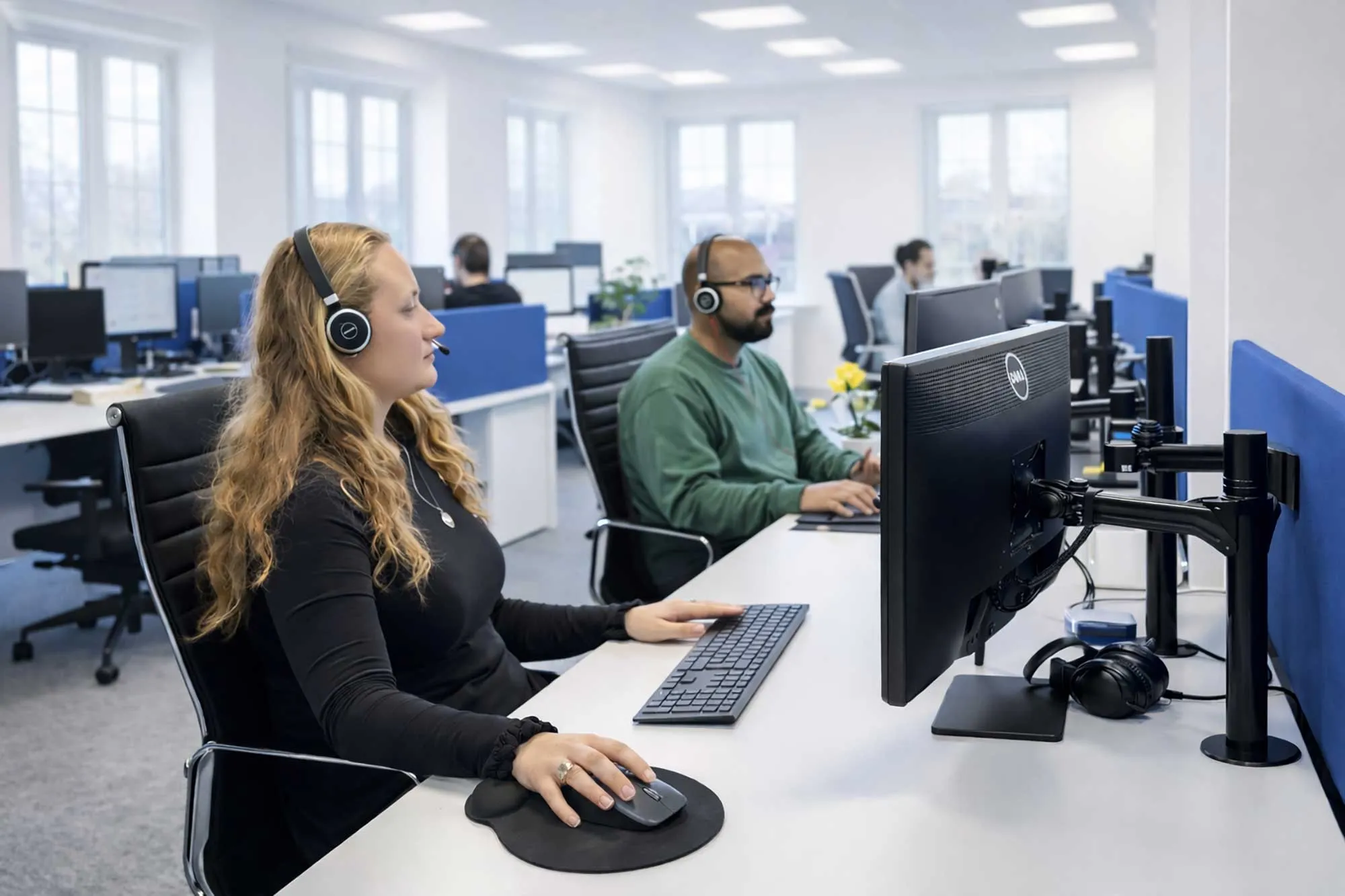 Two office workers wearing headsets and working at desktop computers in a bright, modern open-plan office.