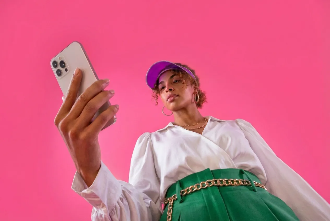 Young woman wearing a purple visor, white blouse, and green pants takes a selfie with a smartphone against a pink background.