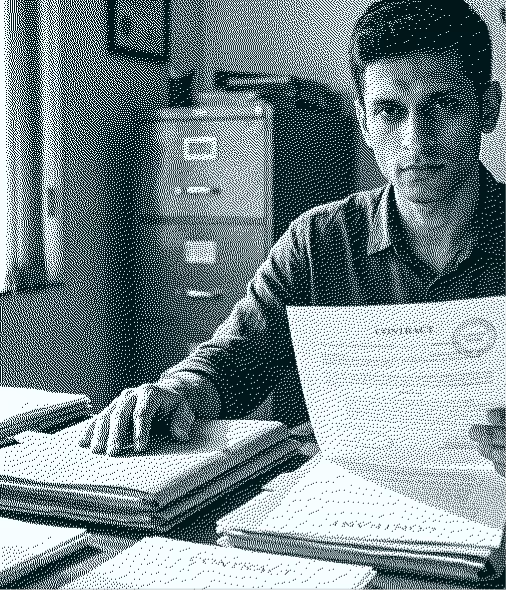 Man sitting at desk holding a document with stacks of papers and a filing cabinet behind him.