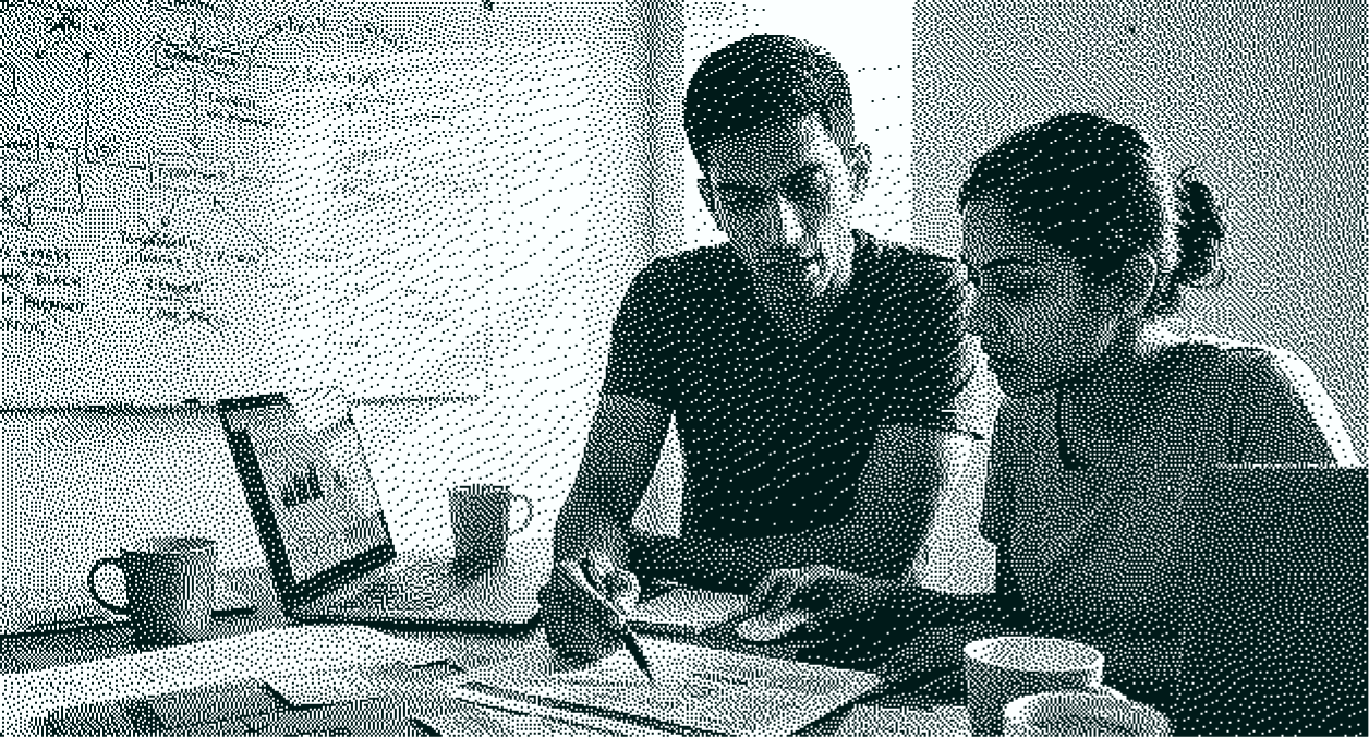 Two people working together at a table with a laptop, documents, and mugs in a bright room.